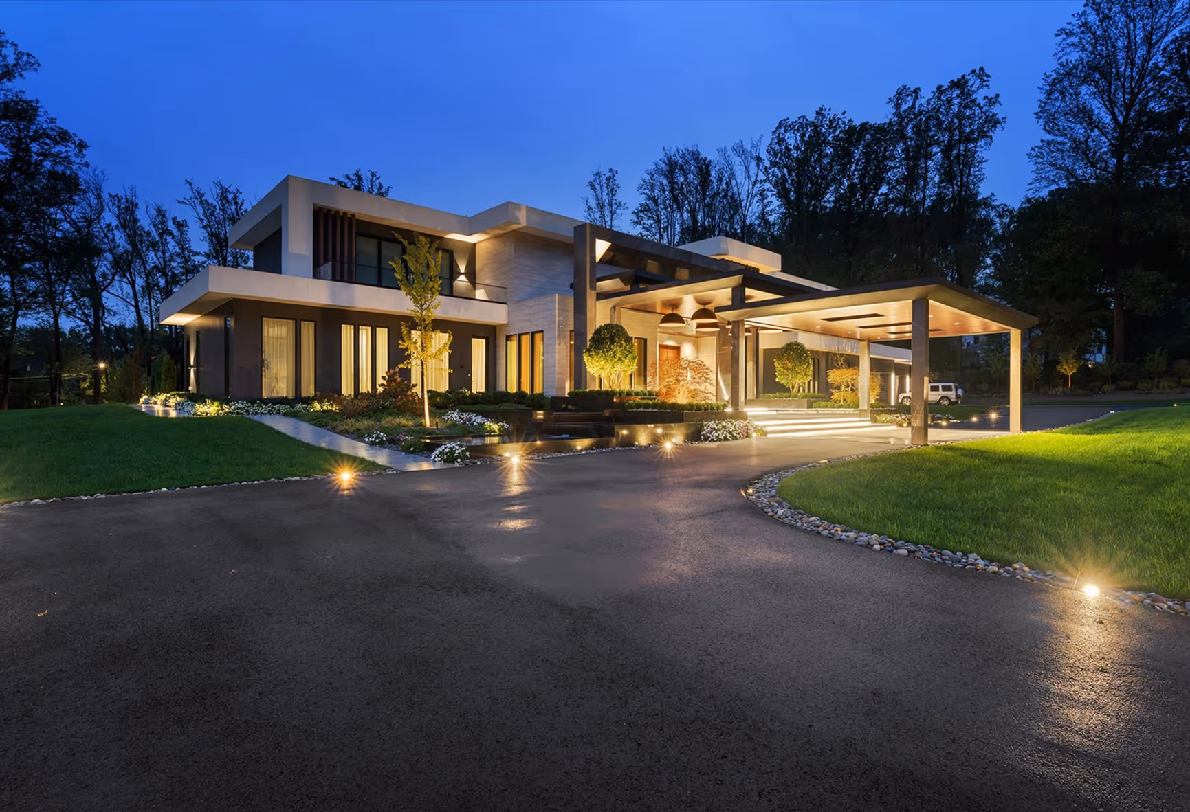 Modern two-story house at dusk with illuminated driveway, landscaped garden, and carport.