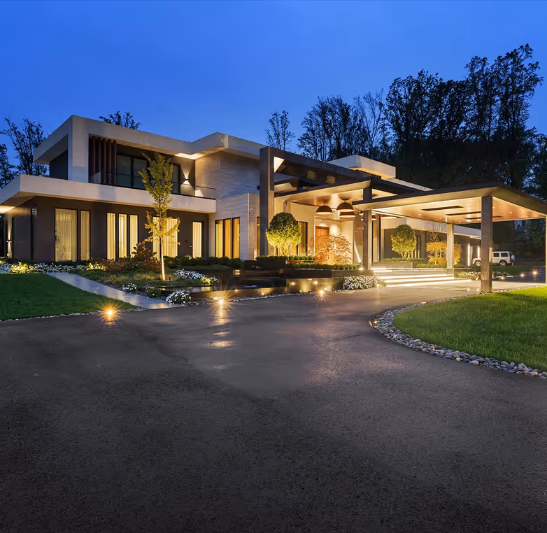 Modern two-story house illuminated at dusk with large driveway, landscaped garden, and covered entrance.