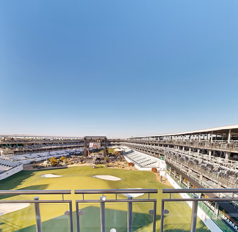 Wide view of an empty golf course surrounded by tiered grandstands under a clear blue sky.