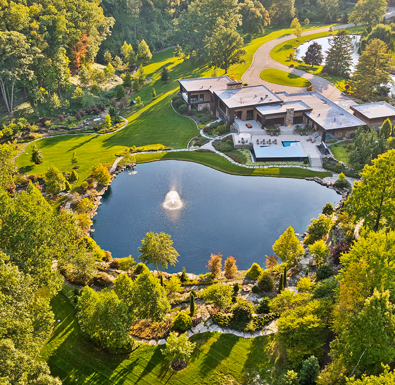Aerial view of a modern house surrounded by lush greenery with a large pond featuring a water fountain in the front yard.