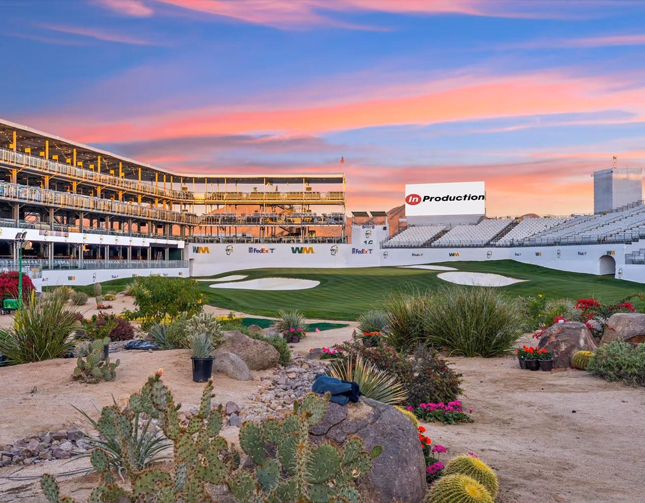 Golf course green and sand bunkers with desert plants in the foreground and empty grandstands under a colorful sunset sky.