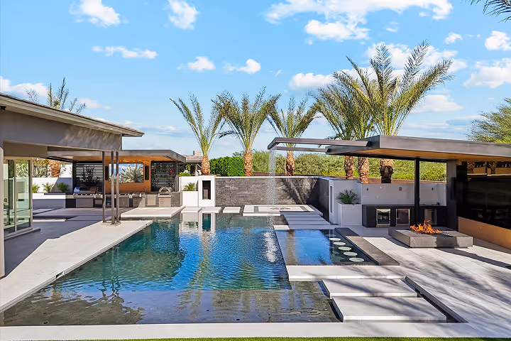 Modern outdoor patio with a rectangular swimming pool, palm trees, and a covered seating area under a blue sky with scattered clouds.