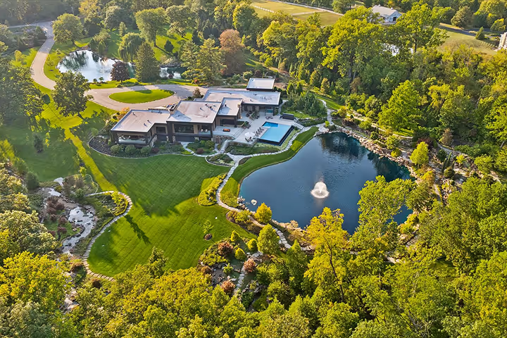 Aerial view of a modern house surrounded by lush green trees, featuring a manicured lawn, a swimming pool, a large pond with a fountain, and winding pathways.