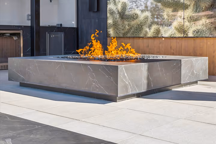 Modern outdoor square fire pit with orange flames on a tiled patio next to a building with glass doors and a large cactus photo on the wall.