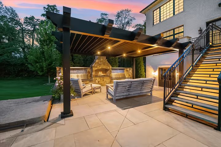 Outdoor patio with wooden pergola, fireplace, cushioned chairs and sofa, next to a white house with illuminated stairs at dusk.