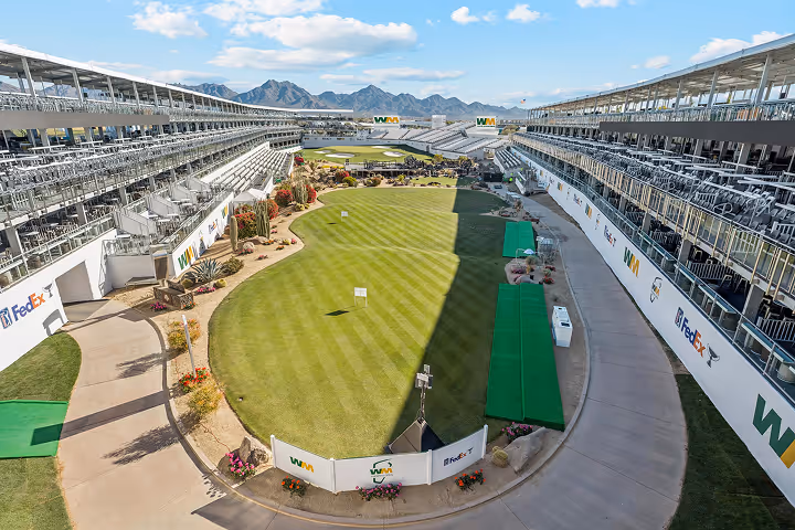 Elevated view of a golf driving range with tiered seating on both sides and mountains in the background under a partly cloudy sky.