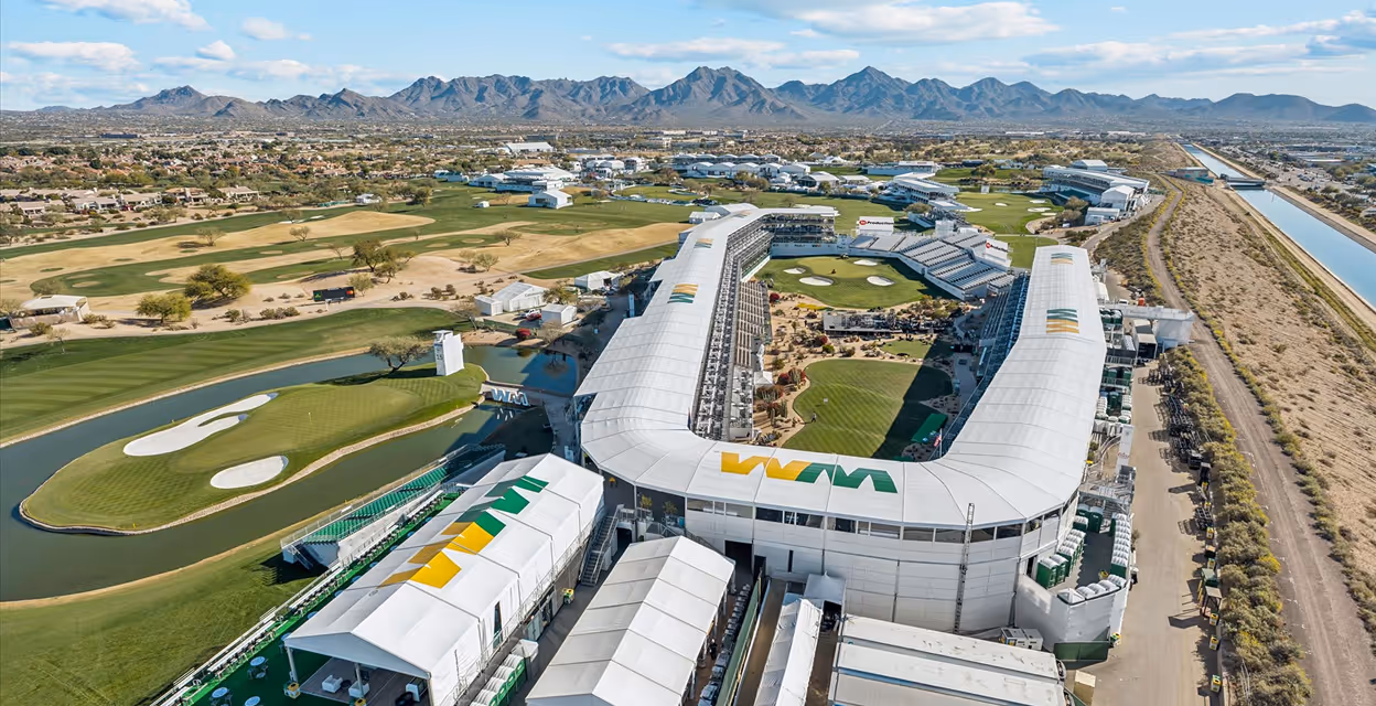 Aerial view of a large golf tournament stadium surrounded by golf course and desert landscape with mountains in the background.