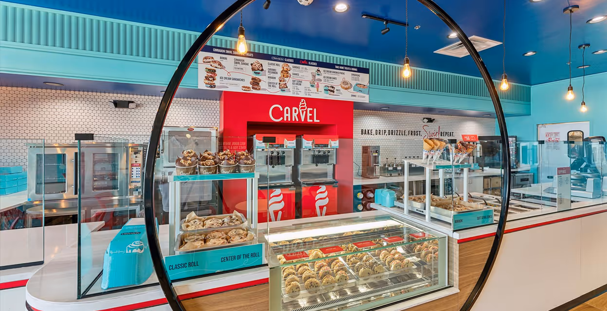 Interior of a Carvel ice cream and pastry shop with display cases of cookies and ice cream machines against a blue ceiling and white tile walls.