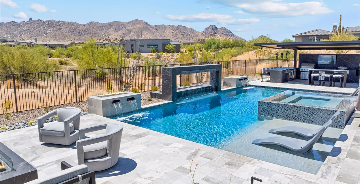 Modern backyard with a rectangular swimming pool, built-in spa, waterfall features, two grey pool loungers submerged in shallow water, and an outdoor kitchen with seating under a pergola.