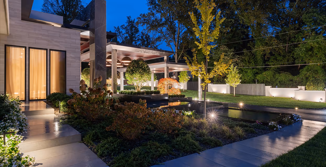 Modern house exterior at dusk with illuminated garden, a water feature, and neatly trimmed bushes and trees along a stone pathway.