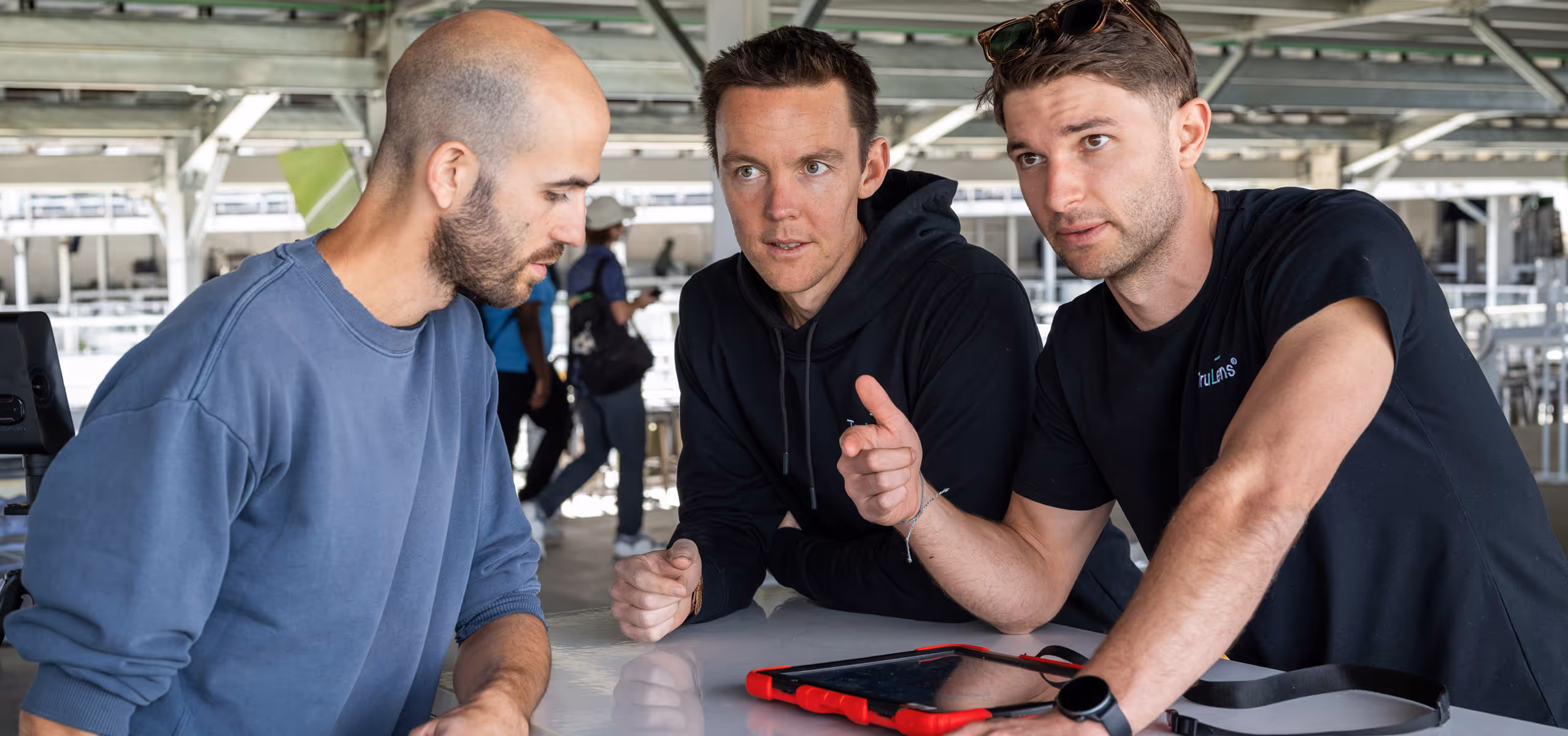 Three men engaged in discussion around a table with a tablet device in an industrial or office setting.
