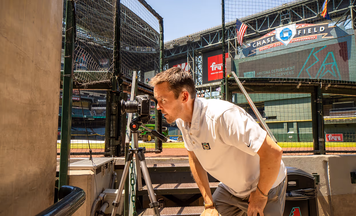 Man in white polo shirt taking a photo with a camera on a tripod inside a baseball stadium near the batting cage.