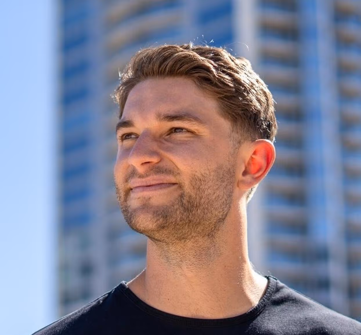 Smiling young man with short brown hair and stubble wearing a black shirt, standing outdoors with a blurred tall modern building in the background.