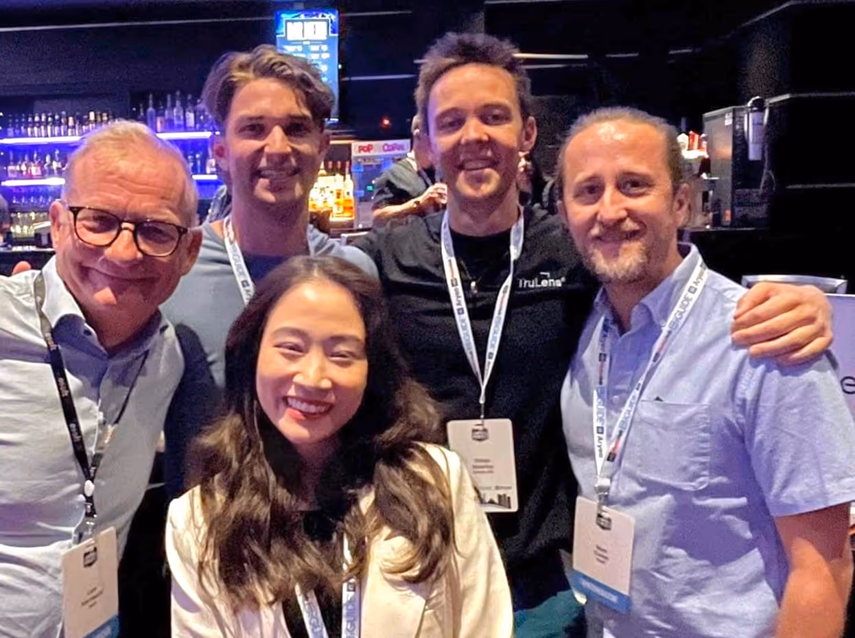 Group of five smiling people wearing conference badges posing indoors at an event.