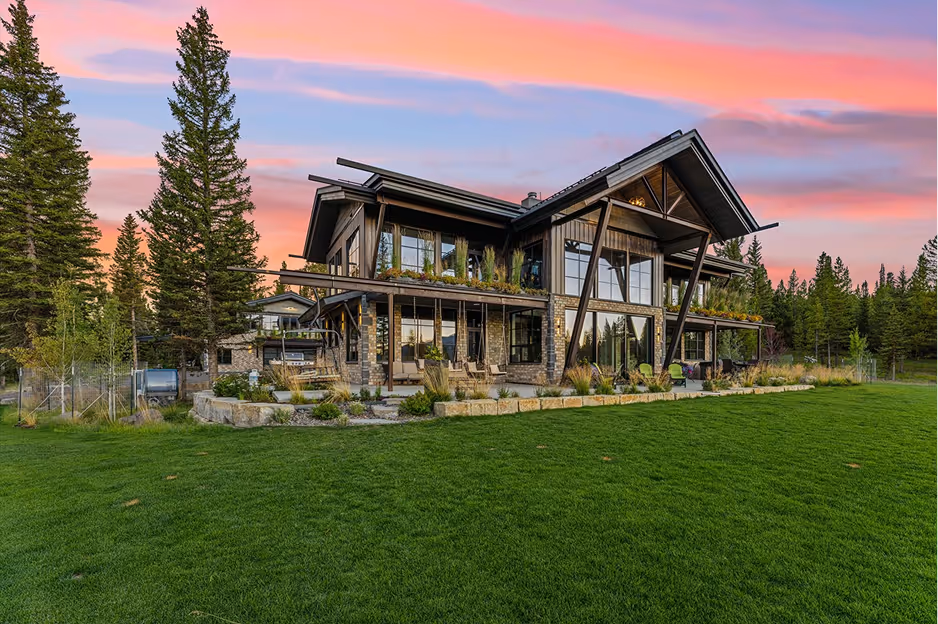 Modern two-story house with large windows, stone and wood exterior, surrounded by green grass and tall pine trees under a colorful sunset sky.