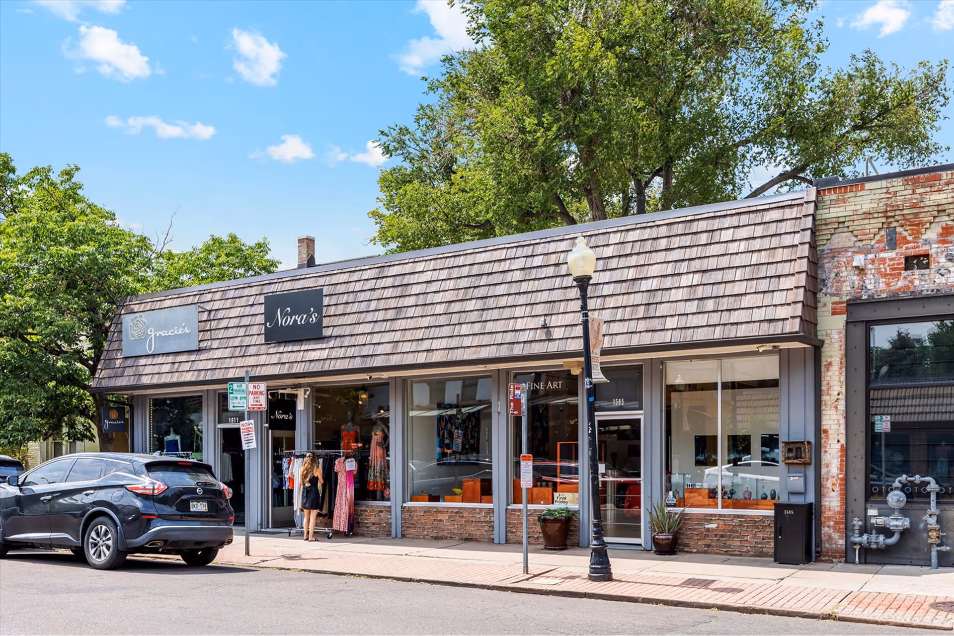 Street view of small retail shops including Gracie's and Nora's with wood shingle roof, a black vehicle parked, and a woman browsing clothing outside.