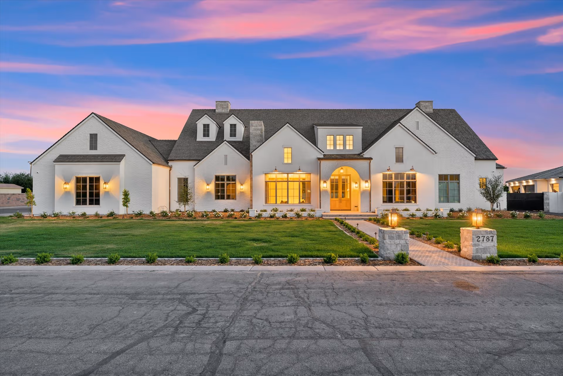 Large white modern farmhouse with gray roof, lit windows and lantern-style exterior lights at sunset.