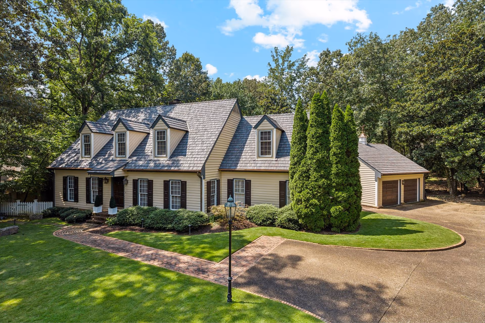 Beige two-story house with dormer windows, a brick walkway, tall shrubs, and a dual garage in a wooded area.