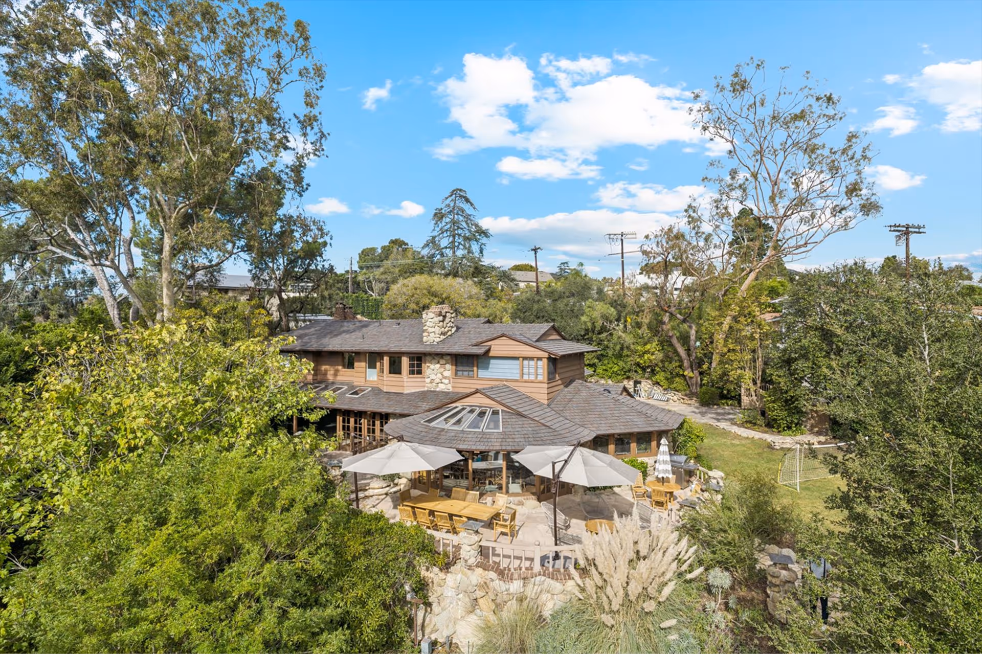 A large, two-story house with stone chimney and wooden siding surrounded by trees, with a patio area featuring umbrellas, tables, and chairs.