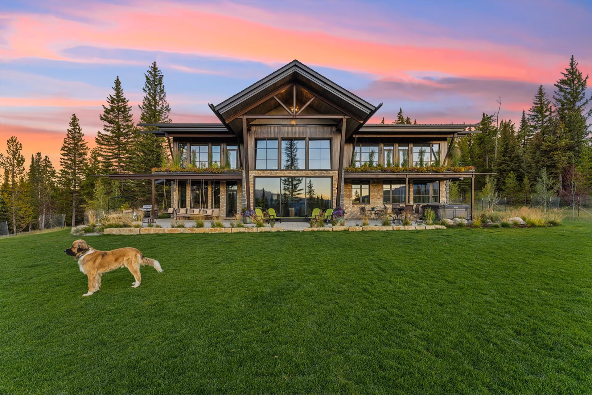 Large modern wood and stone house with tall windows overlooking a green lawn with a dog standing on it at sunset.