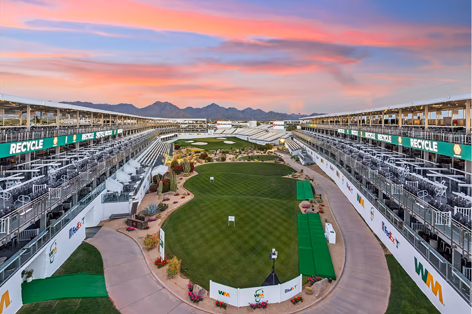 Golf driving range with tiered spectator seating on both sides under a colorful sunset sky.