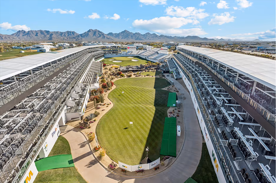 Large outdoor driving range with tiered seating on both sides and mountainous landscape in the background under a partly cloudy sky.