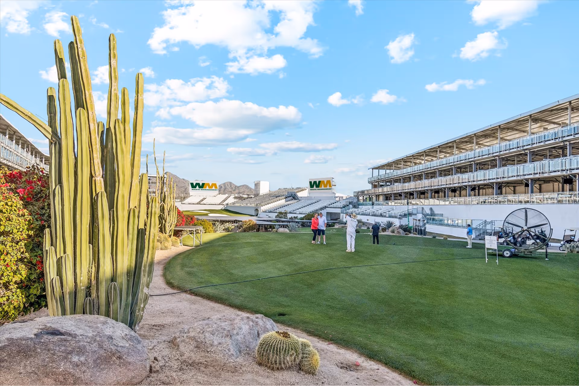 People on a green golf putting area near large cacti with stadium seating and WM Waste Management signs in the background under a blue sky with scattered clouds.