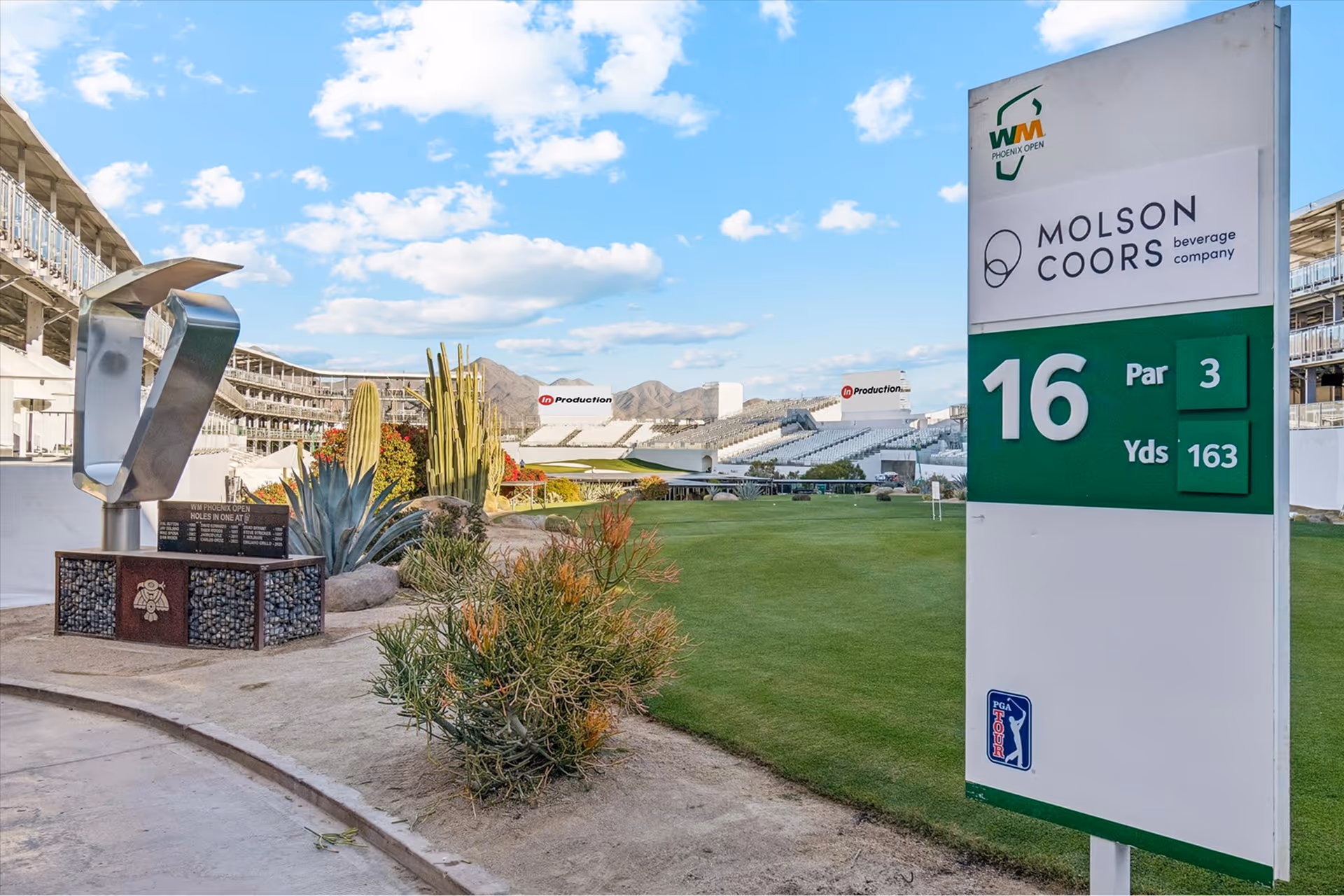 Golf course hole 16 sign with par 3 and 163 yards, trophy on left, desert plants, and stadium seating in background under a partly cloudy sky.