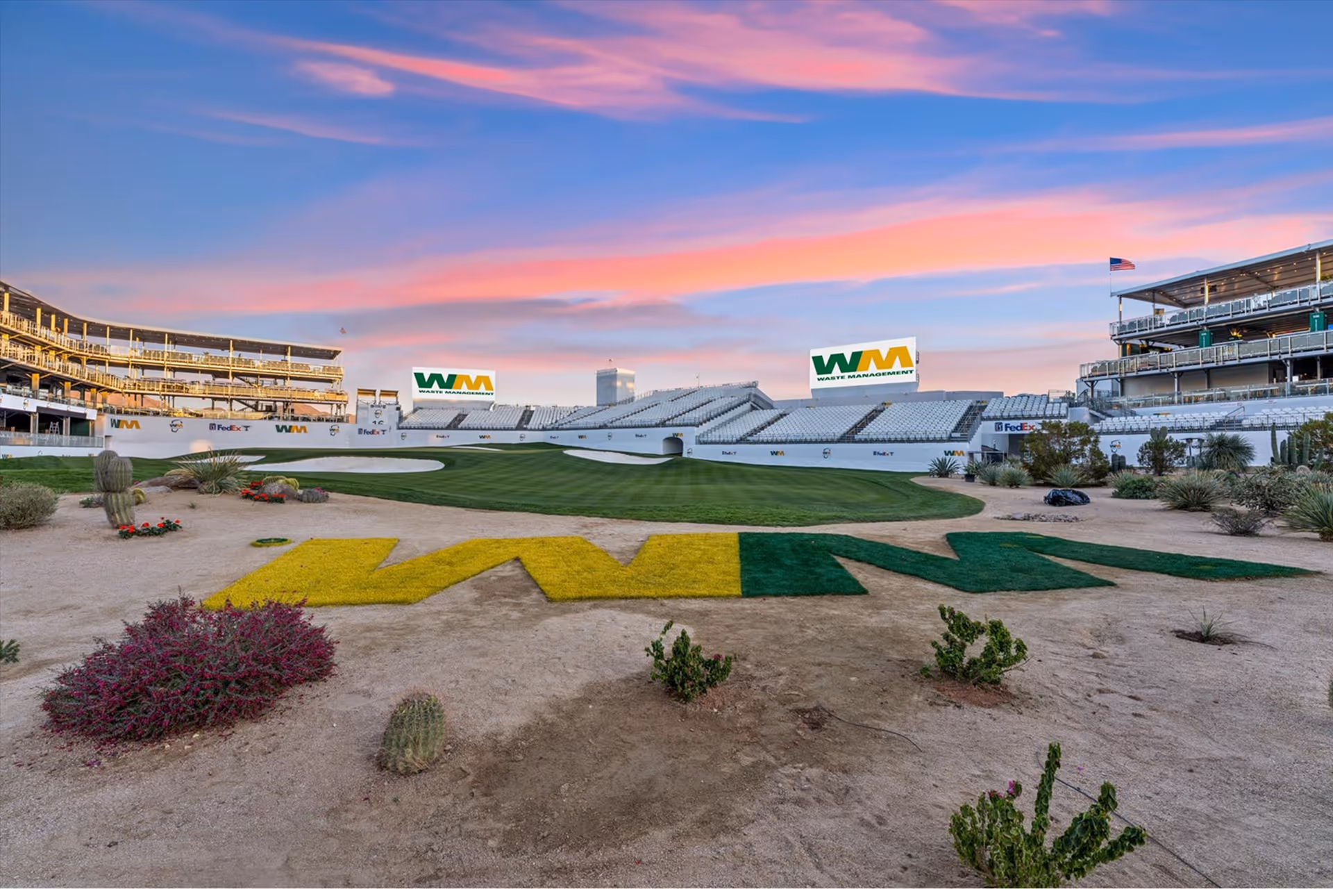 Golf course area with large yellow and green 'WM' logo on sand, grandstands, and a sunset sky with pink clouds.