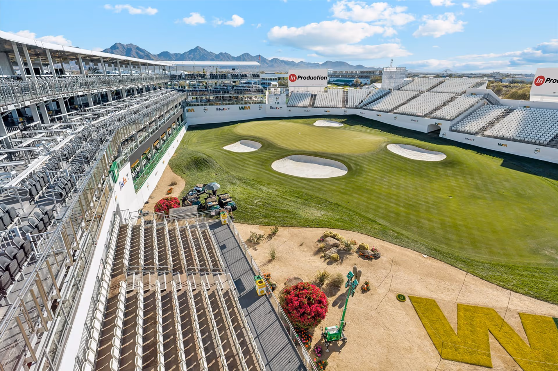 Empty golf course hole surrounded by tiered spectator seating stands under a partly cloudy sky with mountains in the background.