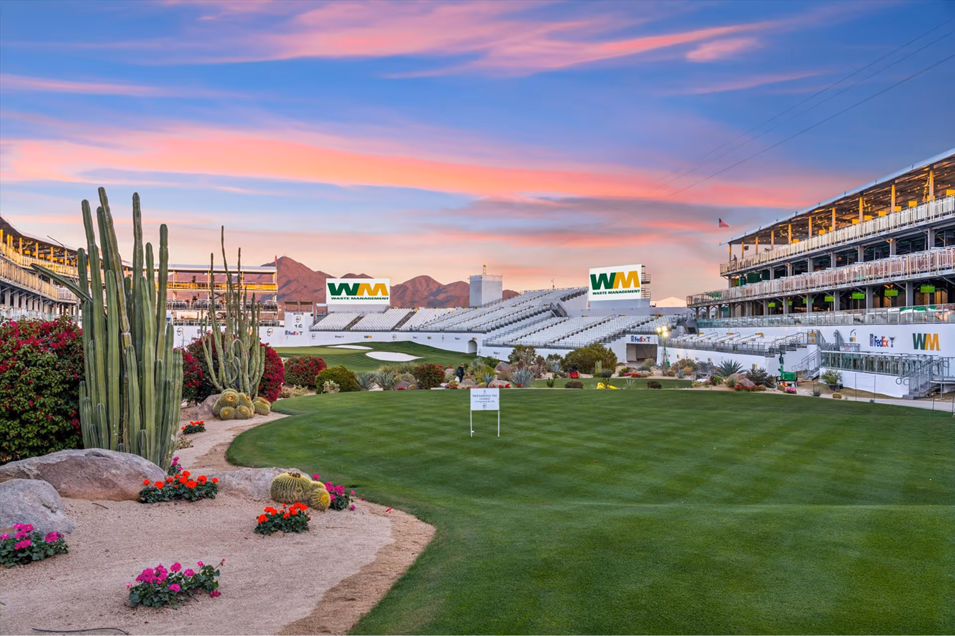 Golf course with green fairway, cacti, and flowers under a colorful sunset sky, with grandstands and Waste Management banners in the background.