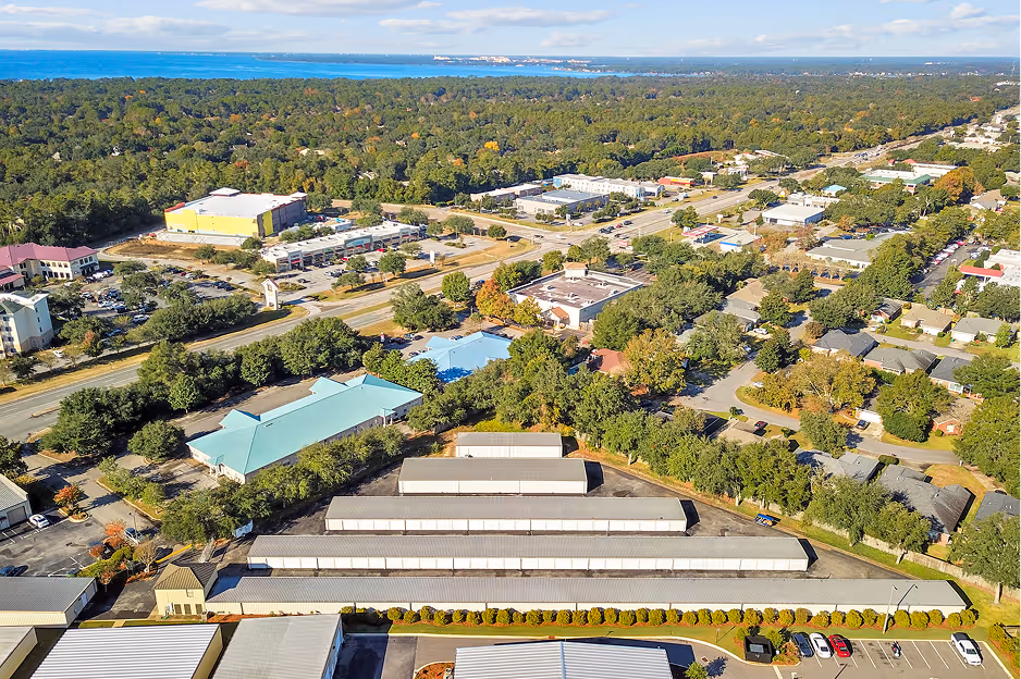Aerial view of a suburban area with rows of long white storage buildings, residential houses, commercial buildings, and a forested area in the distance near a body of water under a cloudy sky.