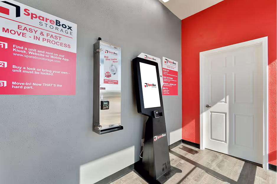 Interior corner of a SpareBox Storage facility with a touchscreen kiosk, a disc lock vending machine, and informational signs on gray and red walls.