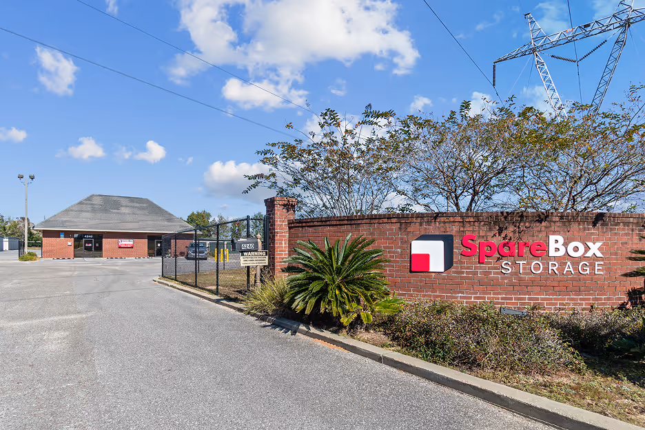 Entrance to SpareBox Storage facility with brick wall sign and gated driveway under a blue sky with clouds.