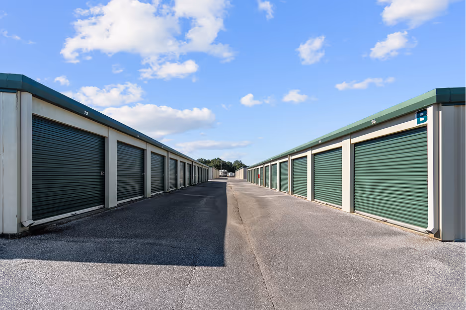 Outdoor storage facility with two rows of closed green roll-up doors under a blue sky with scattered clouds.