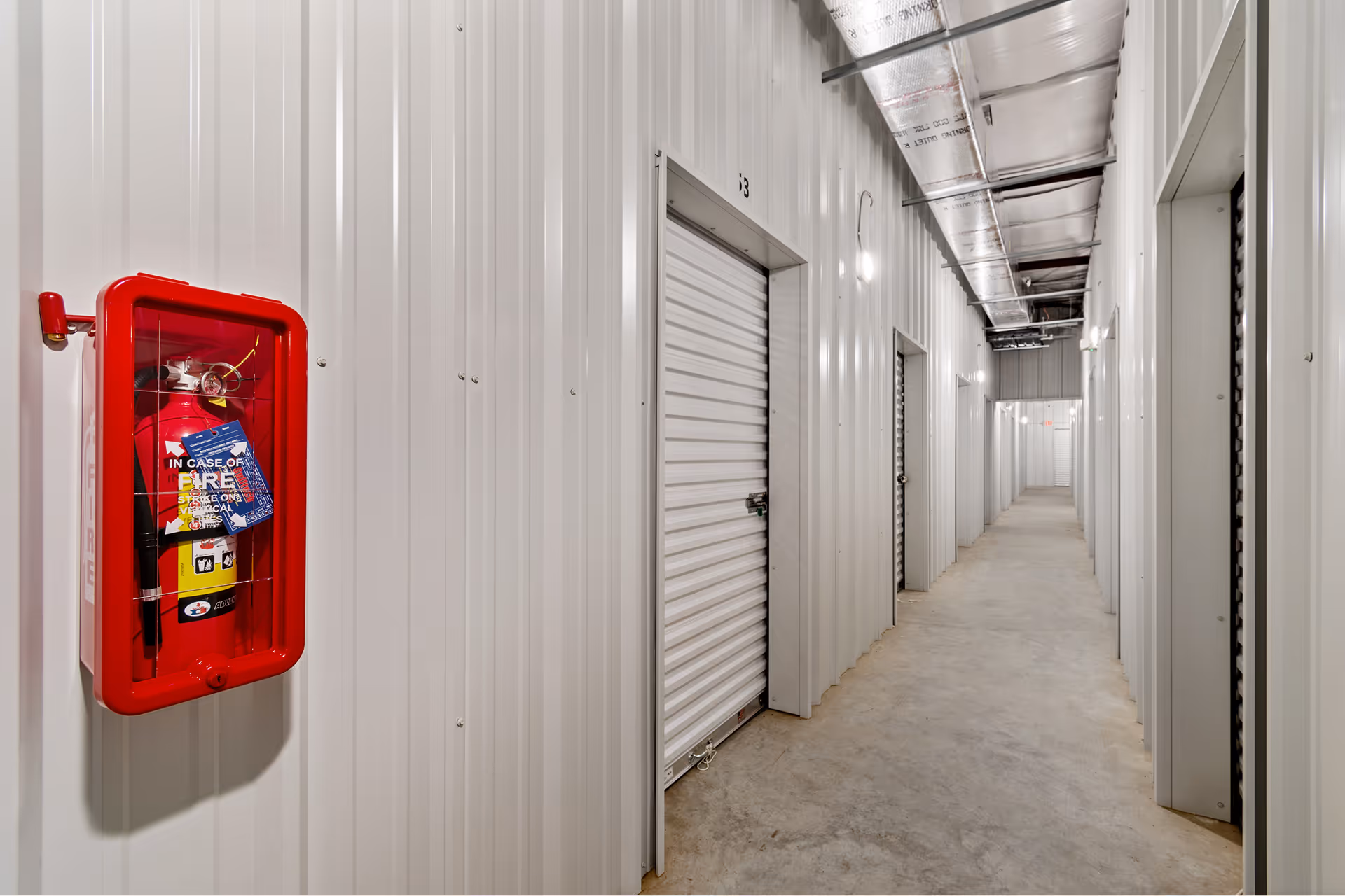 Indoor hallway of a storage facility with white metal roll-up doors and a red fire extinguisher mounted on the wall.