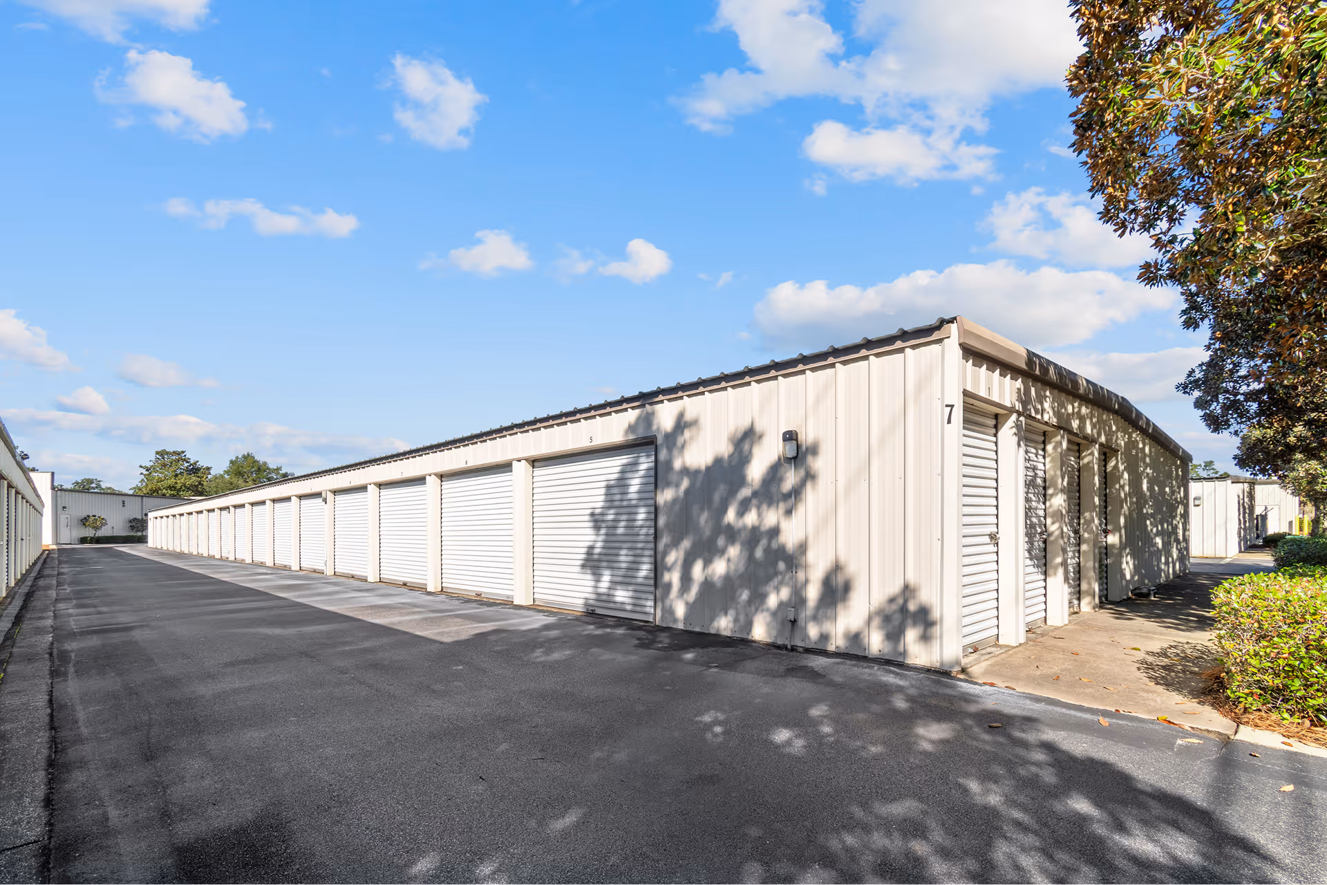 Outdoor storage units with white roll-up doors lining a paved driveway under a blue sky with scattered clouds.