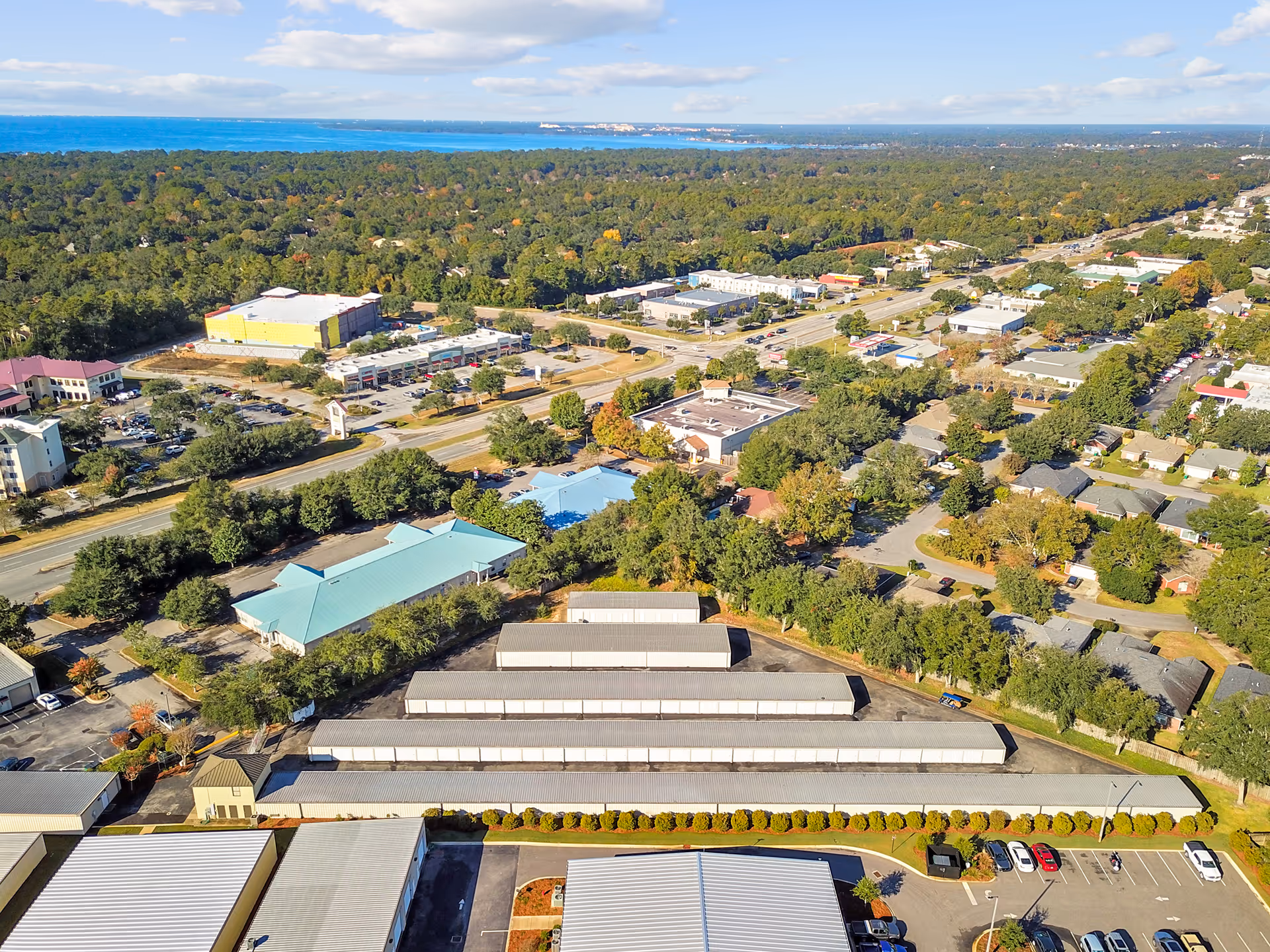 Aerial view of a suburban area with storage units, commercial buildings, scattered trees, and a distant coastline under a partly cloudy sky.