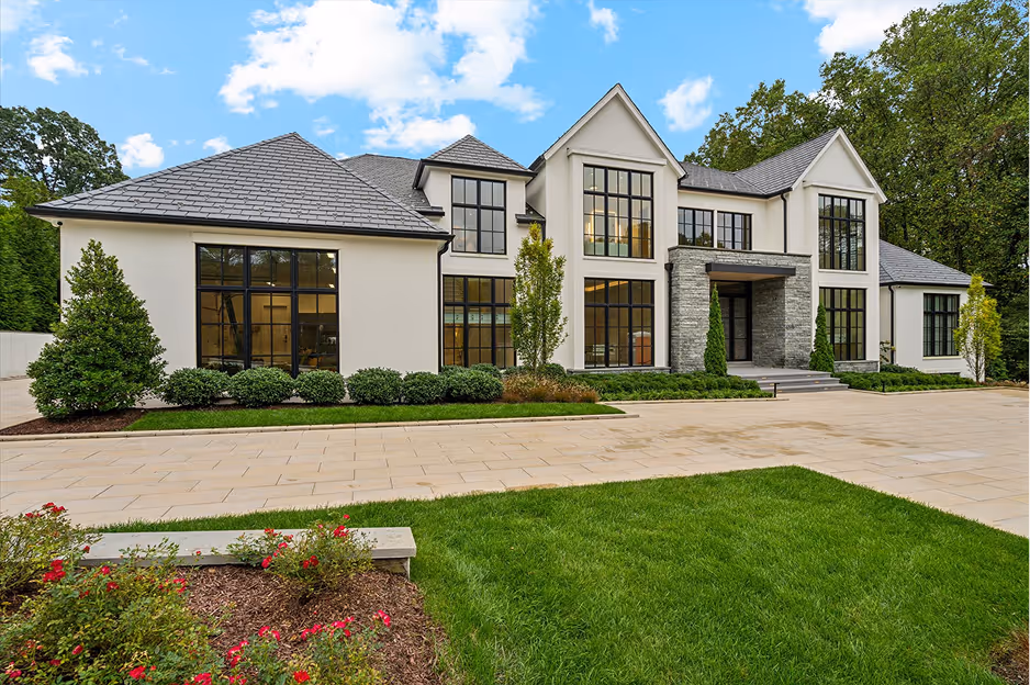 Large modern two-story house with gray roof, multiple tall black-framed windows, stone entrance, and landscaped front yard with green lawn and flowerbeds.