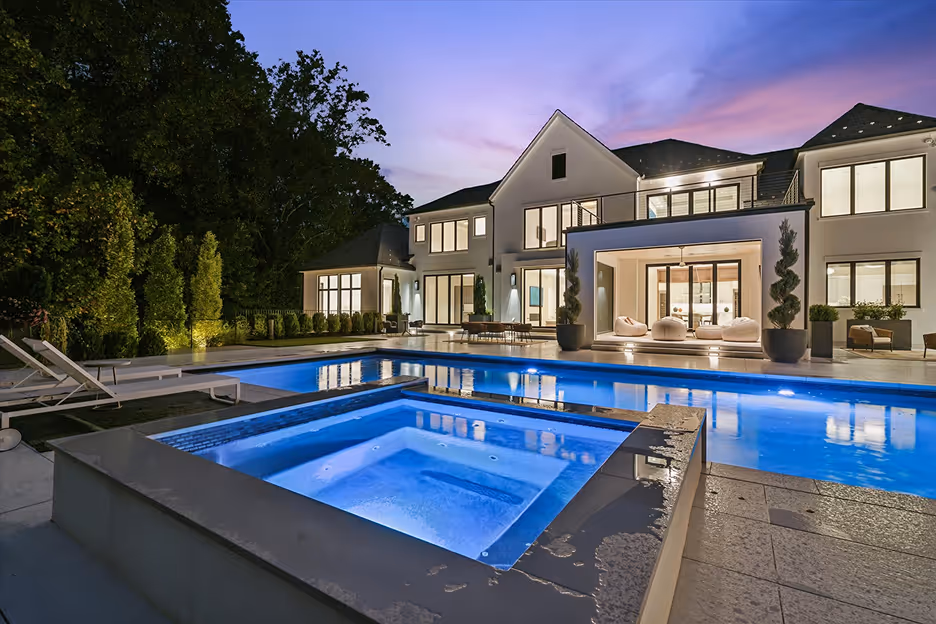 Modern large white house with illuminated rectangular swimming pool and hot tub at dusk.