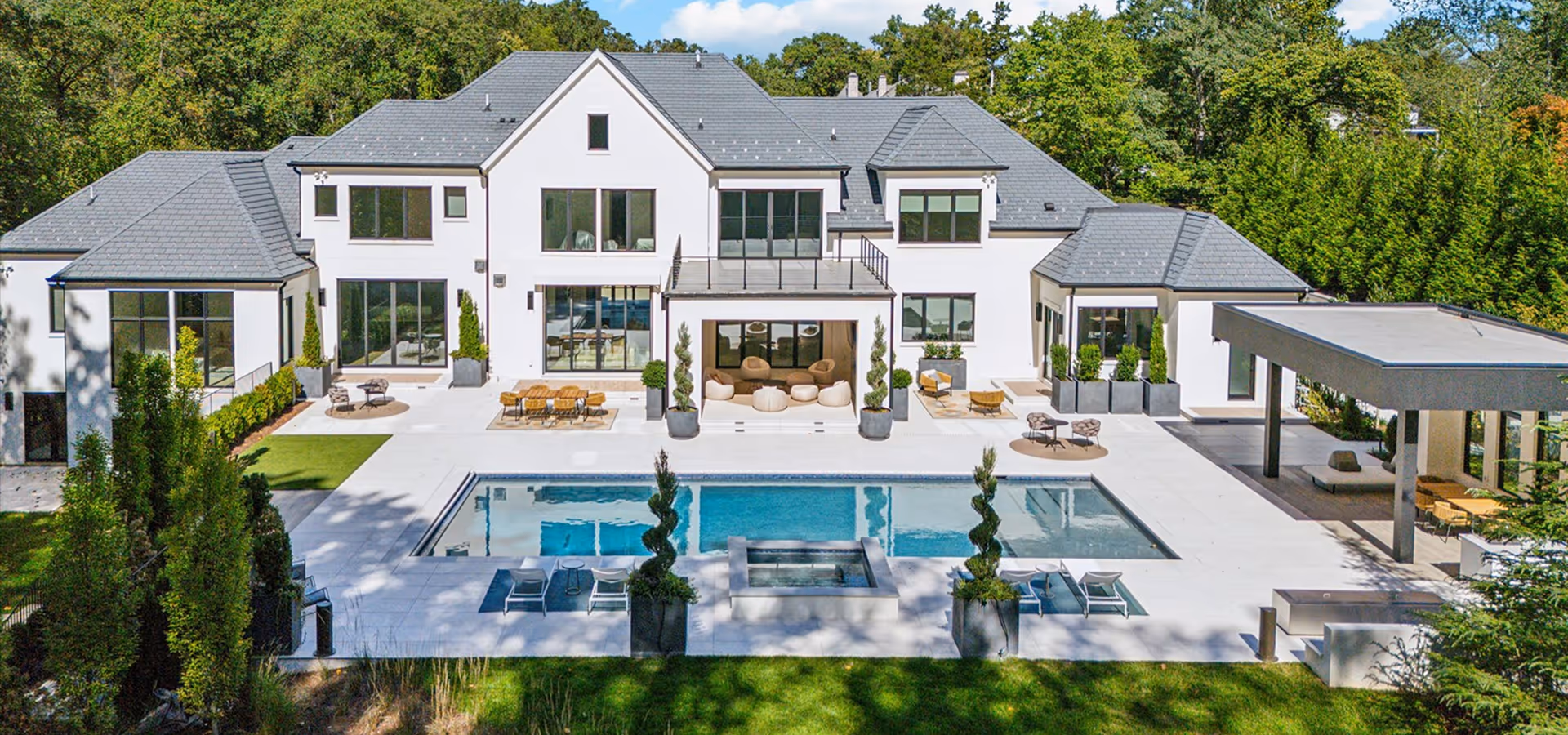 Modern white two-story house with large windows, a central patio with cushioned seating, and a rectangular swimming pool with surrounding lounge chairs and greenery.