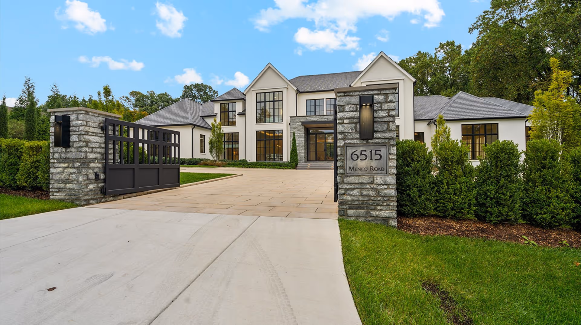 Large modern two-story house with a gated driveway and manicured green hedges and lawn under a blue sky with clouds.
