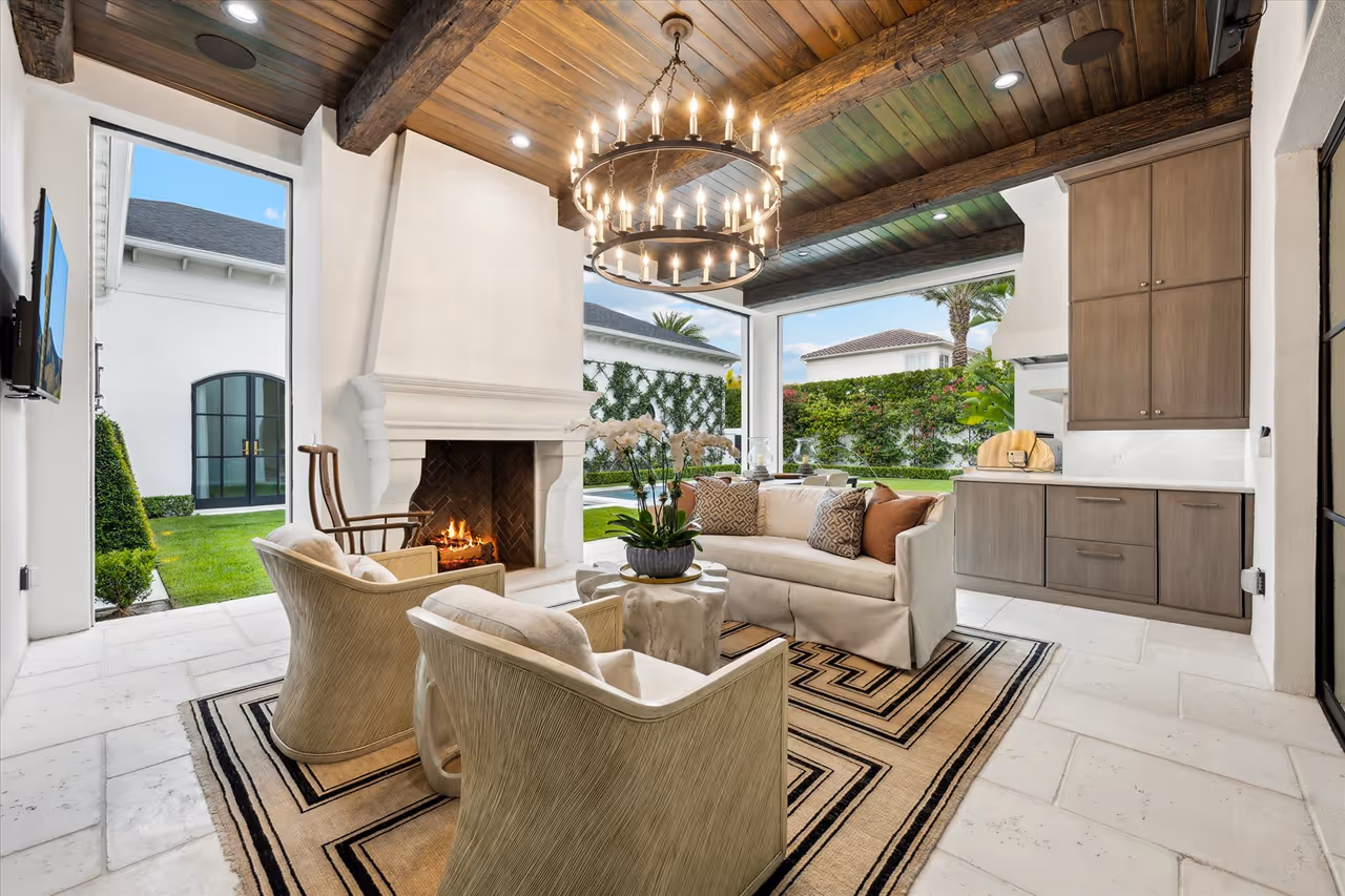 Cozy patio living area with two armchairs, a sofa, a lit fireplace, wooden ceiling, and a view of a green garden outside.