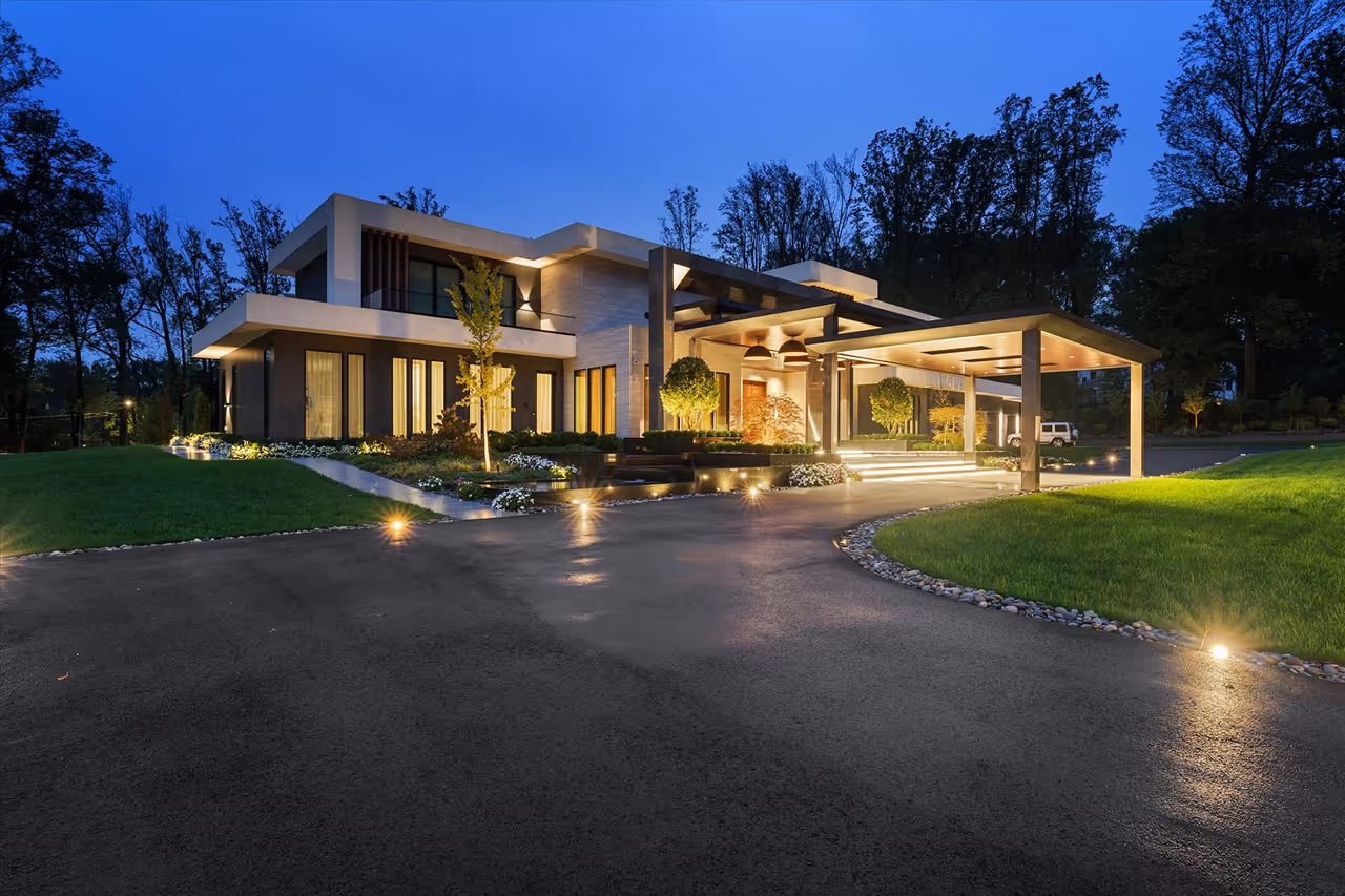 Modern two-story house with outdoor lighting, a driveway, and landscaped garden at dusk.