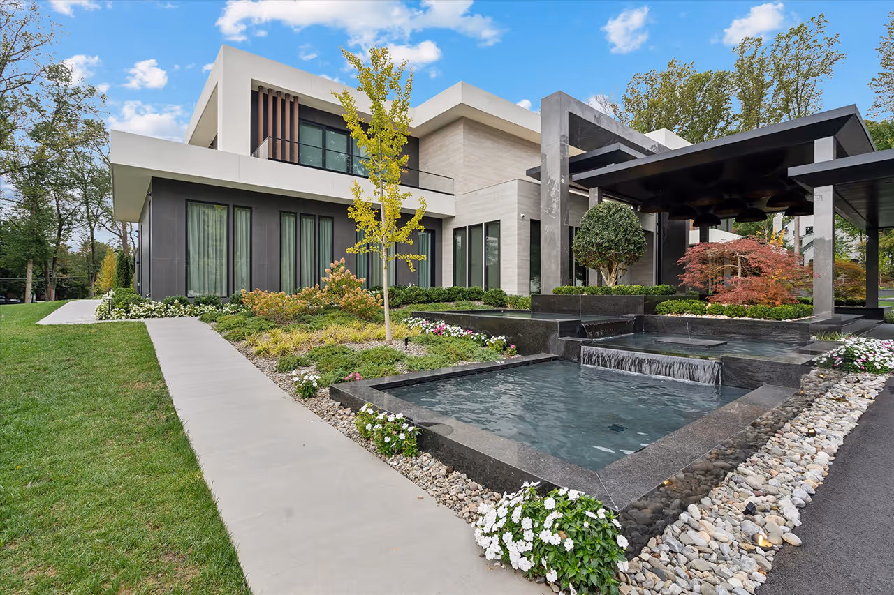 Modern house with a landscaped front yard featuring a small tree, shrubs, flowers, a stone pathway, and a tiered water feature under a dark pergola.