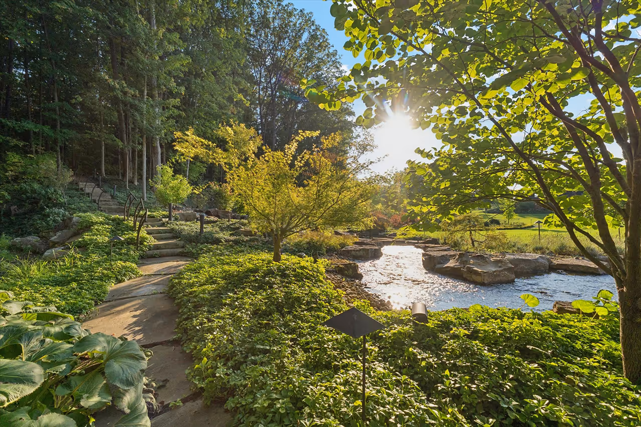 Sunlit garden path surrounded by green shrubs and trees next to a gently flowing rock-edged stream.