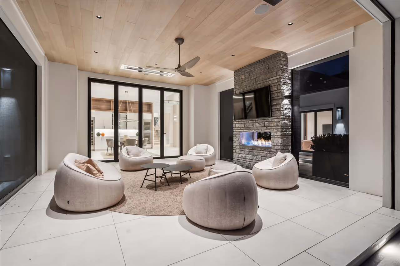 Modern outdoor seating area with six rounded cushioned chairs around a beige rug, two small round tables, a ceiling fan, stone fireplace with TV, and large glass doors.