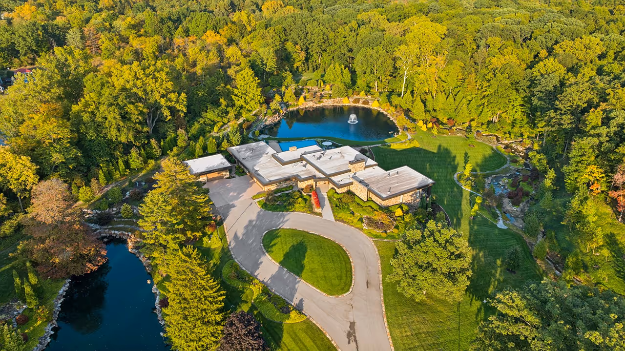 Aerial view of a modern house with white flat roofs surrounded by lush trees, green lawns, a circular driveway, and two decorative ponds, one with a fountain.