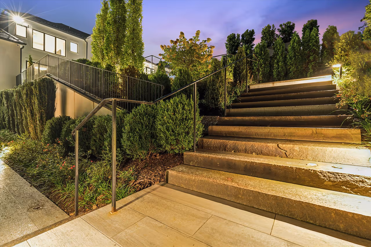 Outdoor stone staircase with metal handrails surrounded by lush greenery and lit by warm lights at twilight.