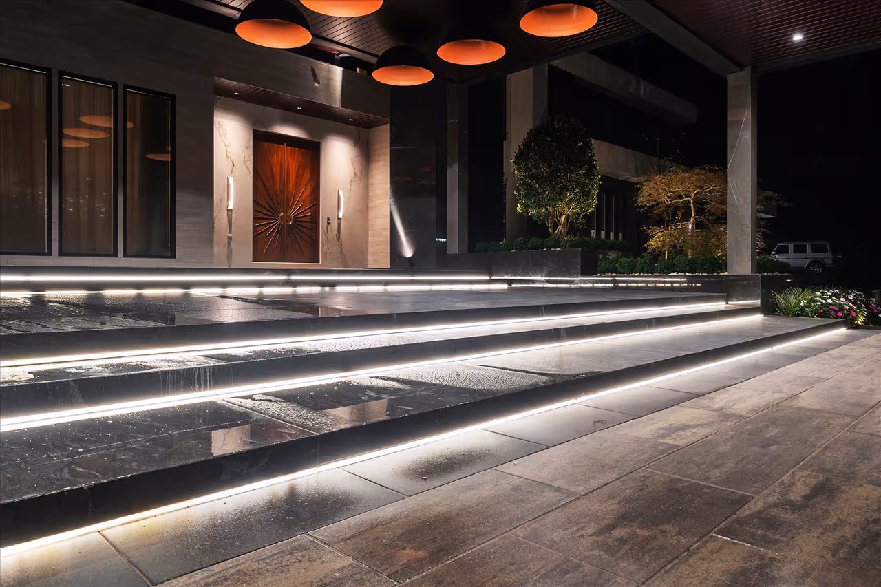 Modern outdoor entryway with illuminated steps, a wooden front door, and surrounding plants at night.
